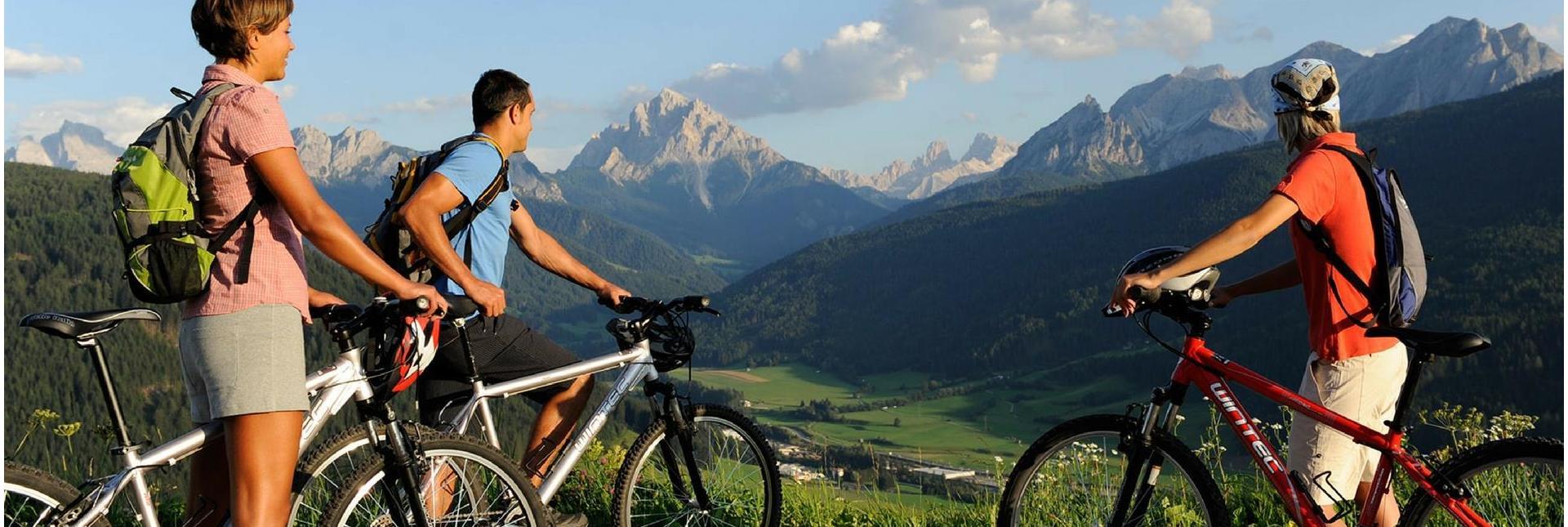 Three cyclists with backpacks look out over a green valley and the Dolomites from a scenic viewpoint.