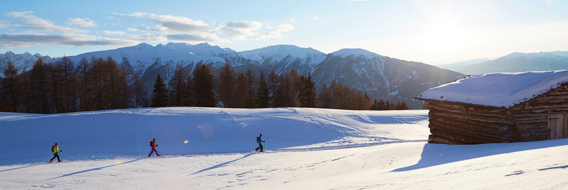 Tre persone fanno escursioni con le ciaspole su un paesaggio innevato con baita e panorama montano.